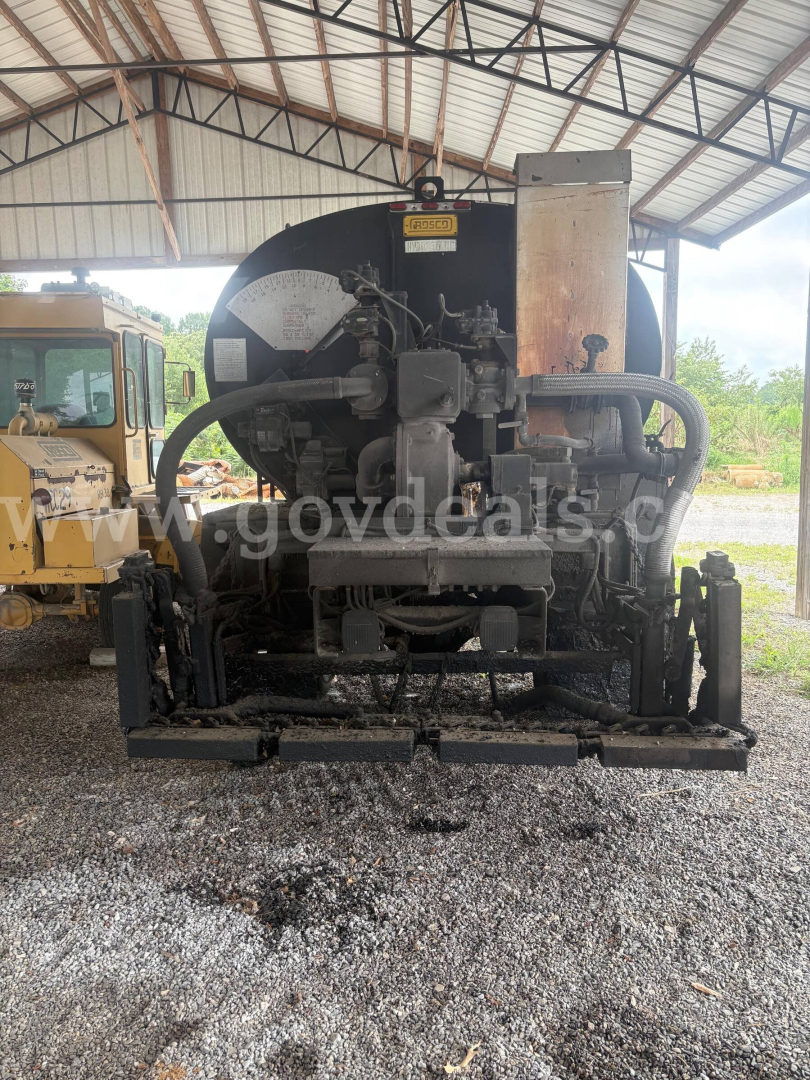 Front view of a black asphalt hot oil truck engine/compartment with hoses, gauges, and metal components under a covered structure.