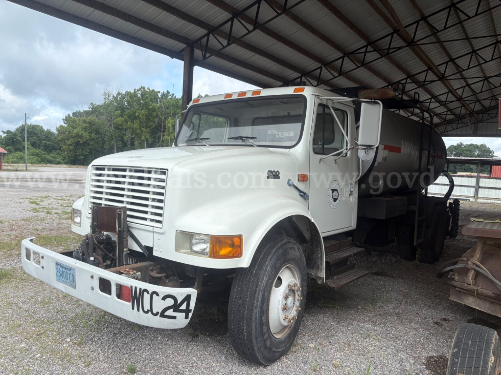 Dump truck chassis with white cab under a metal shelter showing front grille, bumper, and tanker attached; license plate and headlights visible.