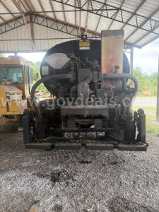 Front view of a black asphalt hot oil truck engine/compartment with hoses, gauges, and metal components under a covered structure.