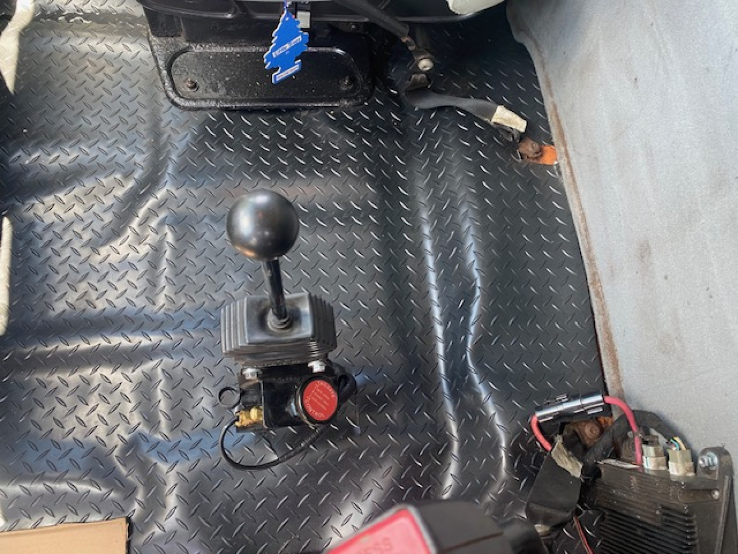 Trucks interior floor showing gear selector with round knob and switch, black diamond-plate floor, wiring, and a red cap valve beside a switch box.