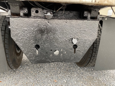 Truck undercarriage with a large textured metal skid plate between dual rear wheels, showing mounting holes and rough surface.