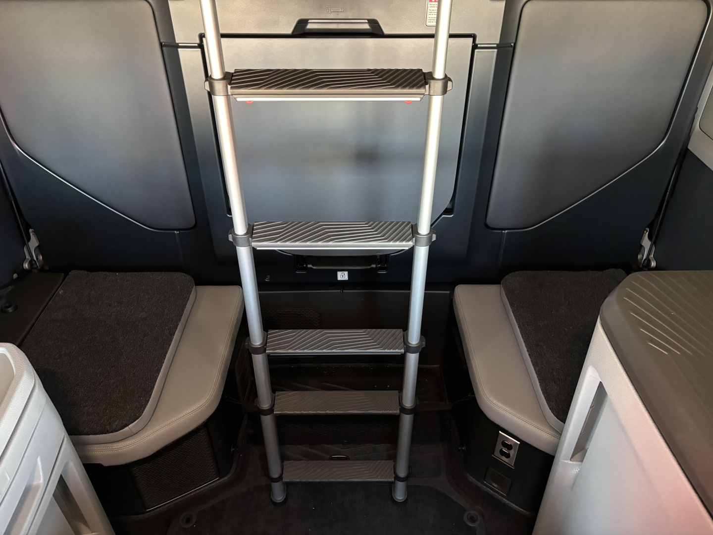 Interior of a Volvo VNL64T860 cab with a foldable metal ladder extending downward from a mezzanine area between the seats; steps with textured treads and metallic frame.