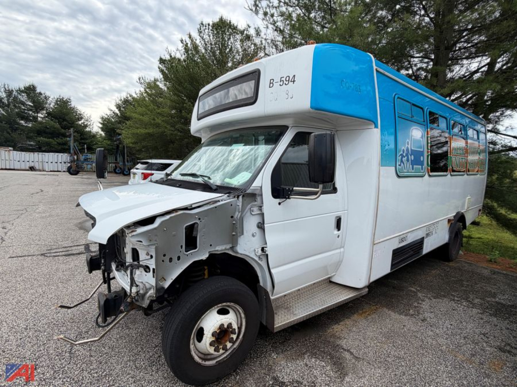 Front left view of a damaged white and blue 2016 Ford E450 shuttle bus. Front hood area is exposed showing wheel, bumper, and headlights removed. Side windows and doors visible with blue accent on upper body.
