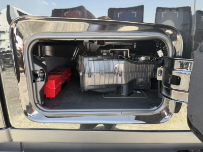 Truck sleeper cab engine bay storage compartment with battery pack on left and large silver air intake housing in center, open rear hatch.