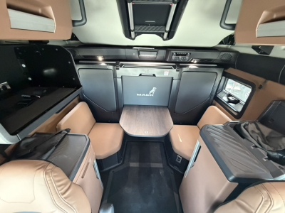Interior of a 2027 Mack PR64T76H sleeper cab showing front seating area, table, and cabinetry with Mack logo on rear cabinet.