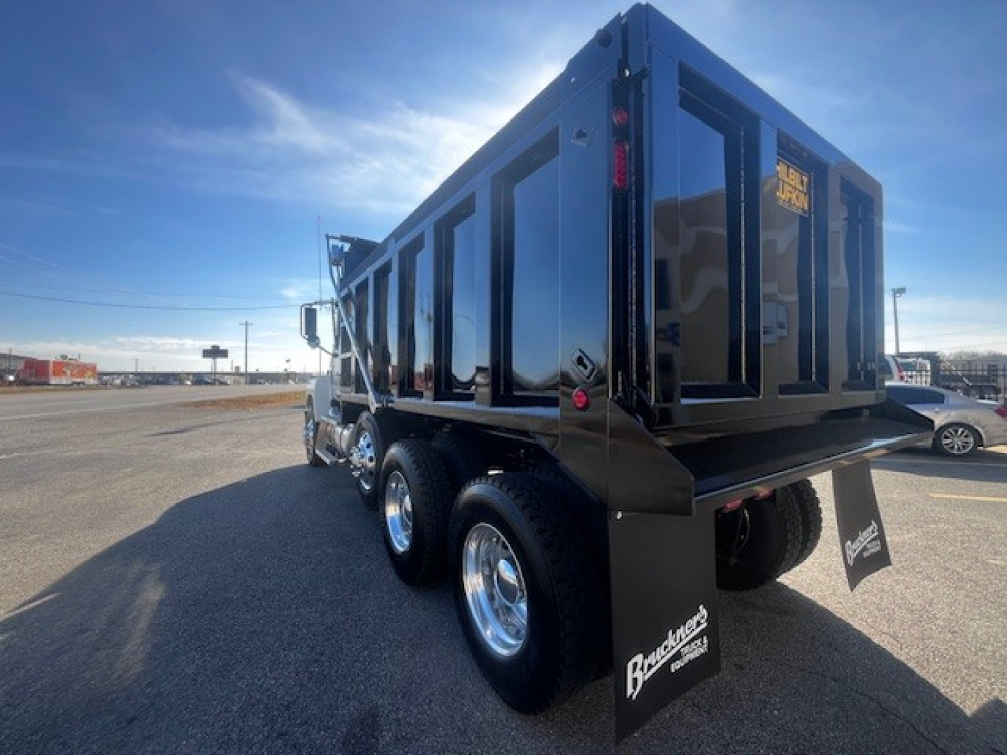 Dump truck rear three-quarter view showing large black dump body on a Mack PI64 with triple rear axles and mud flaps.