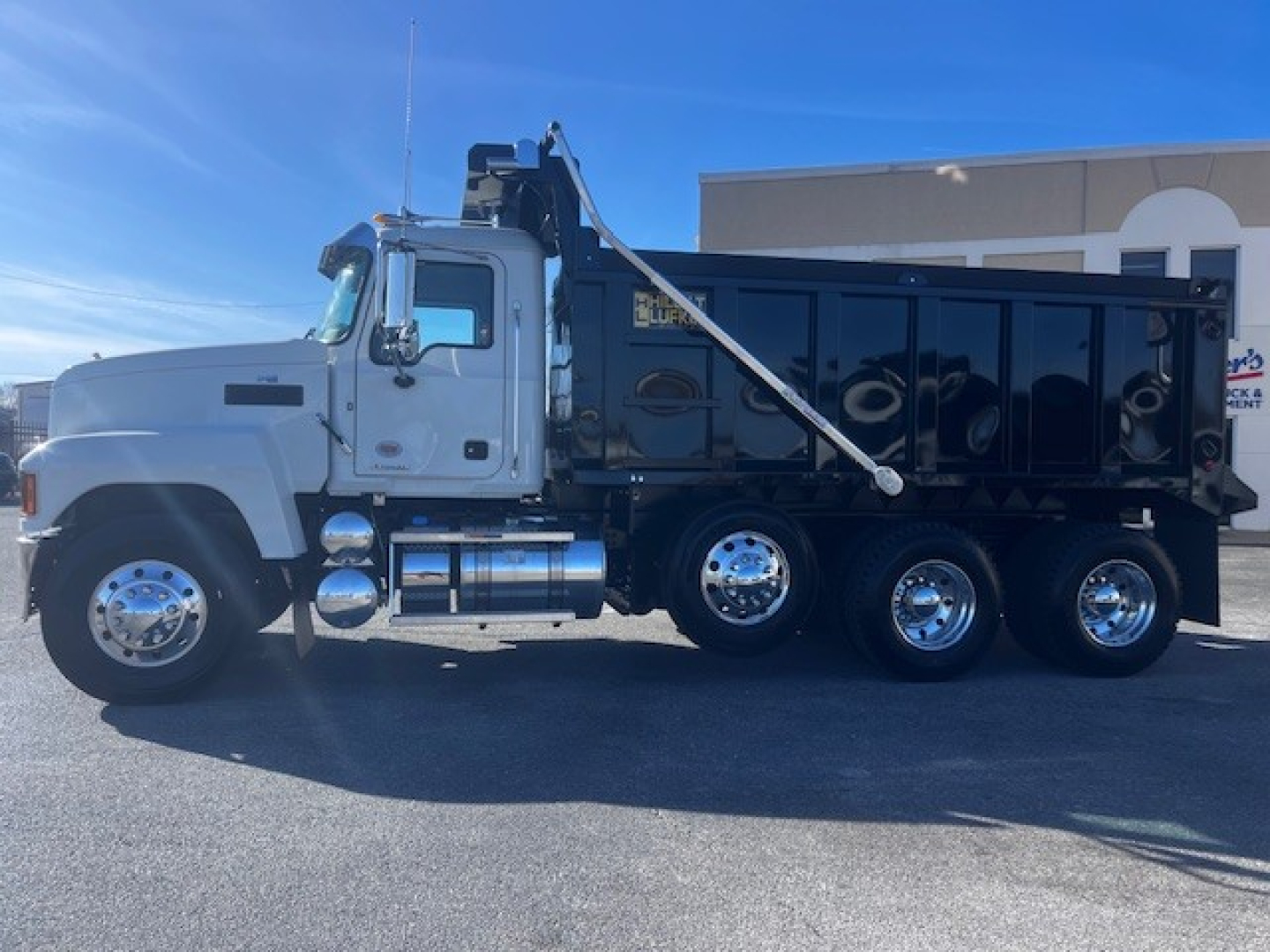 Mack PI64 dump truck side view showing chrome fuel tanks and dump bed with hydraulic hoist system.