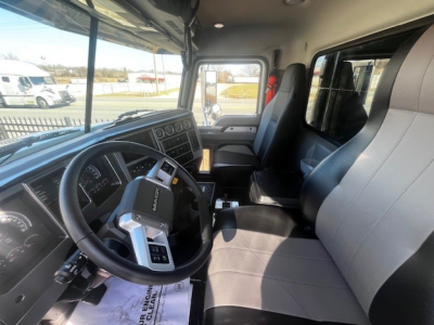 Interior cab view of a 2026 Mack PI64 dump truck showing steering wheel with controls, dashboard gauges, black and gray seats, door, and side window.