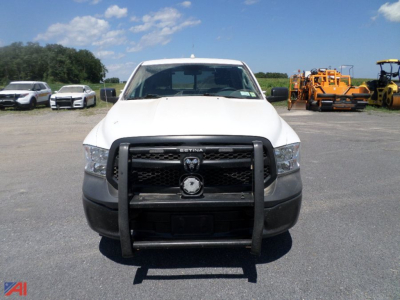 Ram 1500 Crew Cab pickup truck front view with grille guard and Ram emblem, white body, in a lot with other vehicles and construction equipment in background.