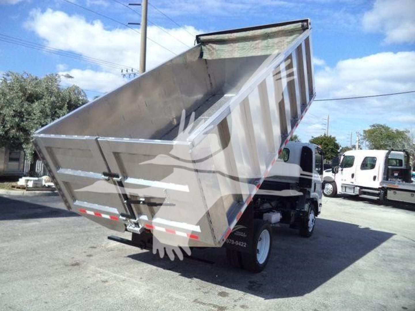 Dump bed of a 2017 Isuzu NPR HD dump truck, tilted up to show cargo bed and hydraulic mechanism, side view with other trucks in background.