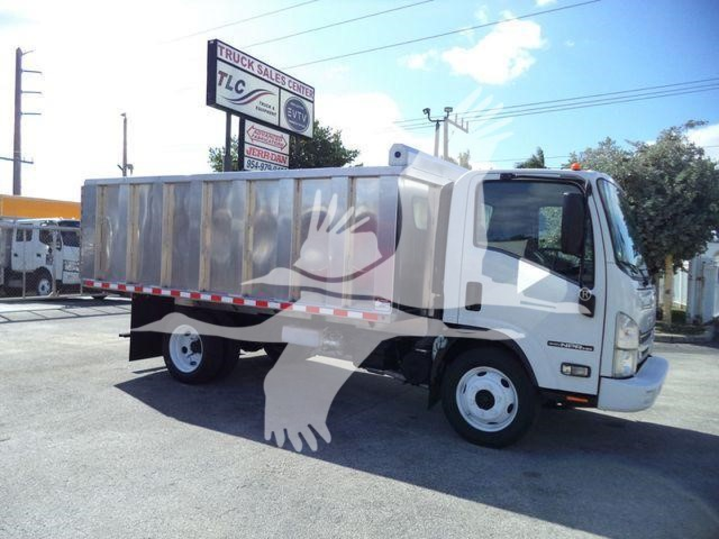 Isuzu NPR HD dump truck in a dealership lot, side view with silver cargo bed and white cab.
