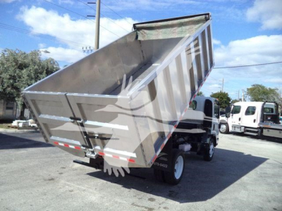 Dump bed of a 2017 Isuzu NPR HD dump truck, tilted up to show cargo bed and hydraulic mechanism, side view with other trucks in background.