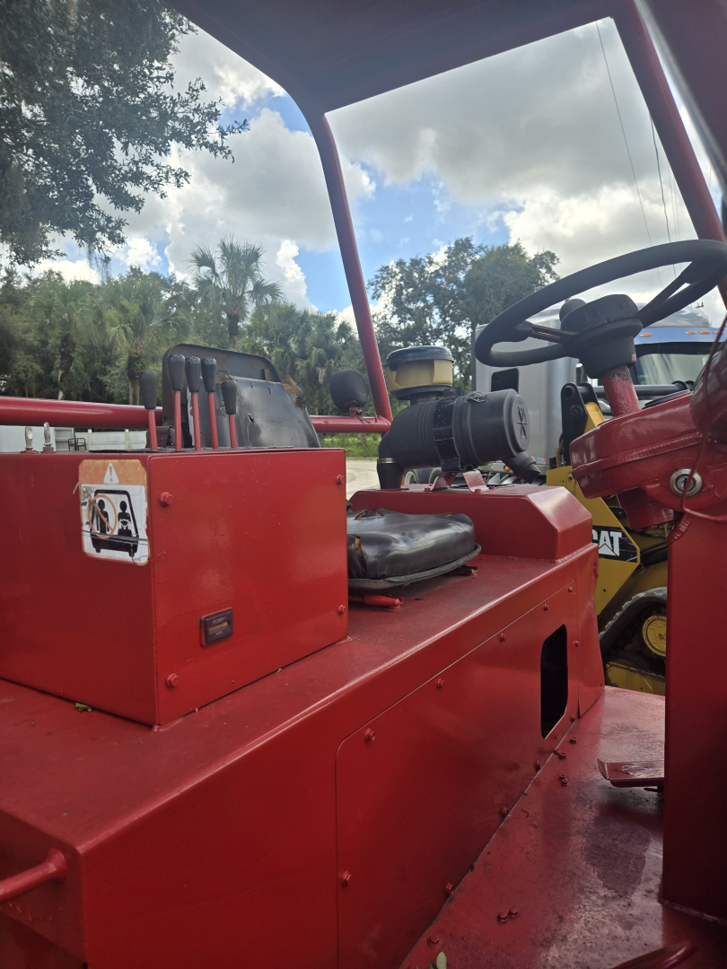 Equipment view of a red telescopic lift truck with operator seat, steering wheel, and control panels; visible engine area and warning label on side.
