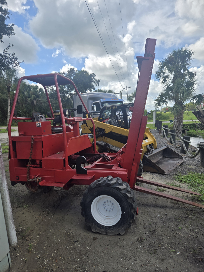 Red telescoping lift with vertical strut raised, attached to front, and white rear wheel, outdoor lot with other machinery in background
