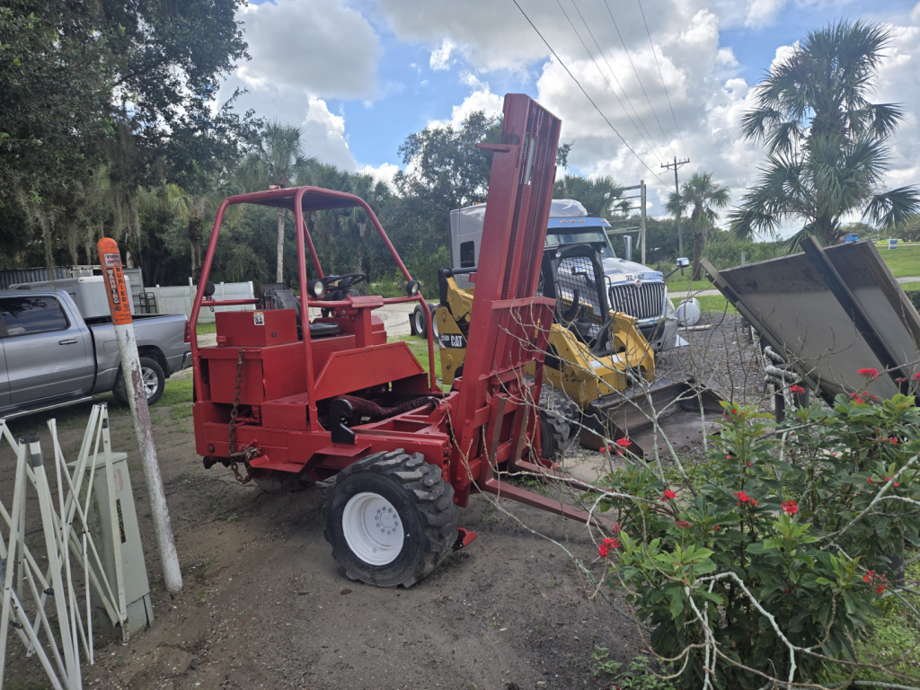 Lift equipment side view showing a red telescopic piggyback forklift with vertical lifting arm and white rear wheel, parked on dirt with other machines in background.