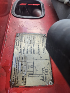 Plate on red telescopic lift showing specifications: wheelbase, capacity, model, serial, attachments; label with diagrams and text, screws at corners.