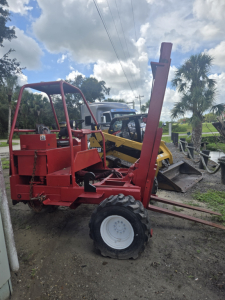 Red telescoping lift with vertical strut raised, attached to front, and white rear wheel, outdoor lot with other machinery in background