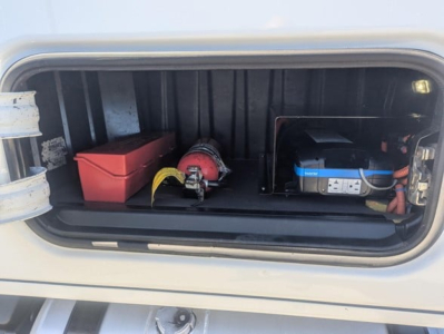 Truck battery and storage area showing a red toolbox, a red circular air hose reel, a blue-labeled battery, and wiring in a Freightliner Cascadia engine bay storage compartment.
