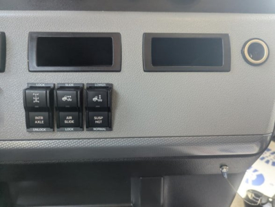Dashboard panel with multiple black toggle switches labeled LOCK, INTR AXLE, ABR SLIDE, SUSP HLT, LOWER, NORMAL; two empty rectangular slots and a circular cigarette lighter style outlet on Freightliner Cascadia dashboard.