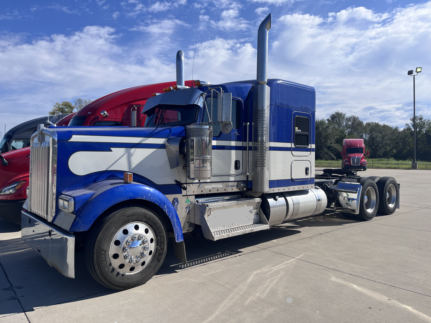 Kenworth W900L 2023 conventional sleeper truck in blue and white livery, front hood with chrome bumper, dual exhaust stacks, side steps, and large front wheel.