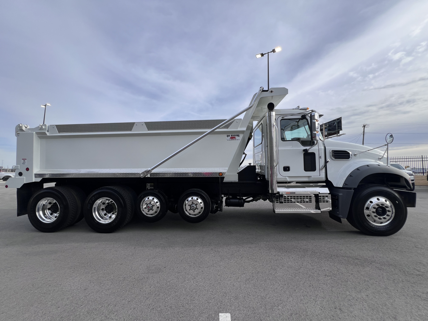 Dump truck side view showing Mack GR64F dump body with 6-wheel tandem rear, chrome wheels, and cab-mounted steps. No visible logos or text legible beyond truck design.