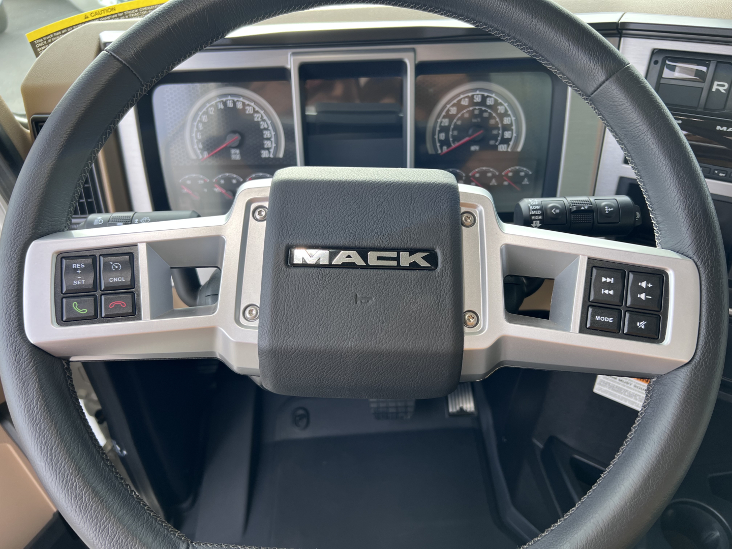 Steering wheel of a Mack GR64F truck showing the Mack logo center and control buttons on both sides; dashboard gauges visible in the background.
