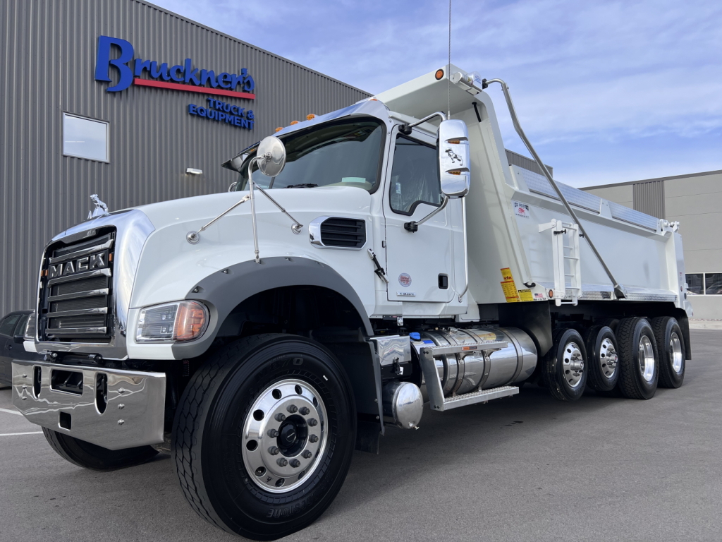 Mack GR64F dump truck in a showroom lot with chrome bumper and dump bed, side view of cab showing mirrors and exhaust.