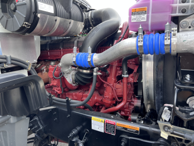 Mack GR64F engine bay showing red engine block, large turbo hoses, and purple coolant reservoir with blue hose clamps.