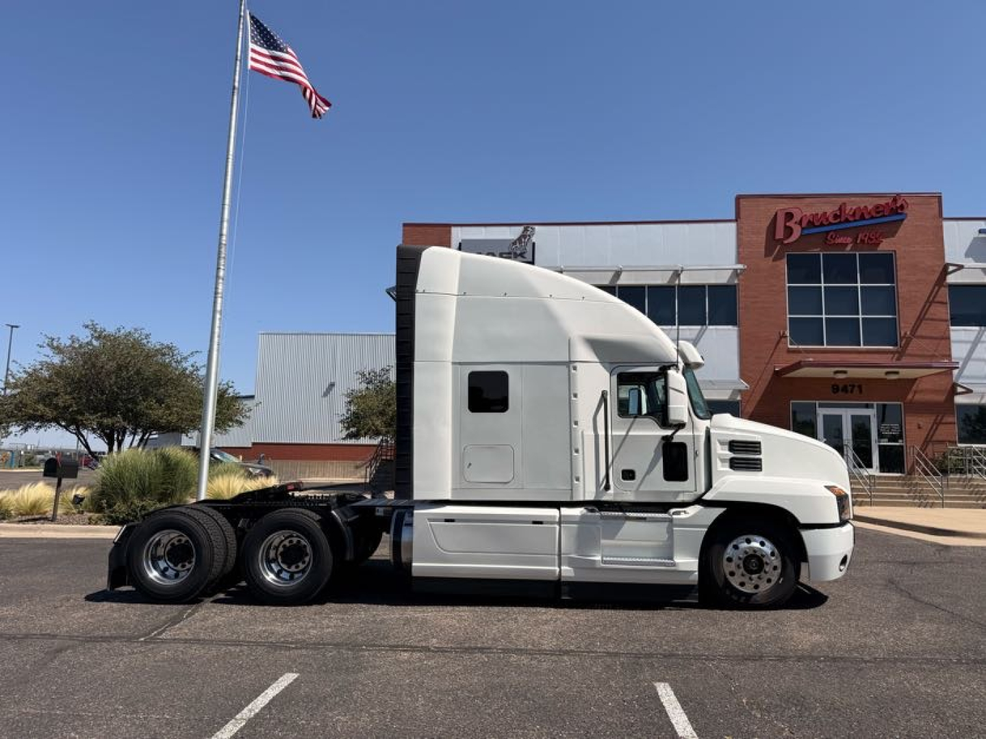 Truck side view of a white 2020 Mack ANTHEM semi-truck in a parking lot, sleeper cab, three rear axles, adjacent to a building with American flag.