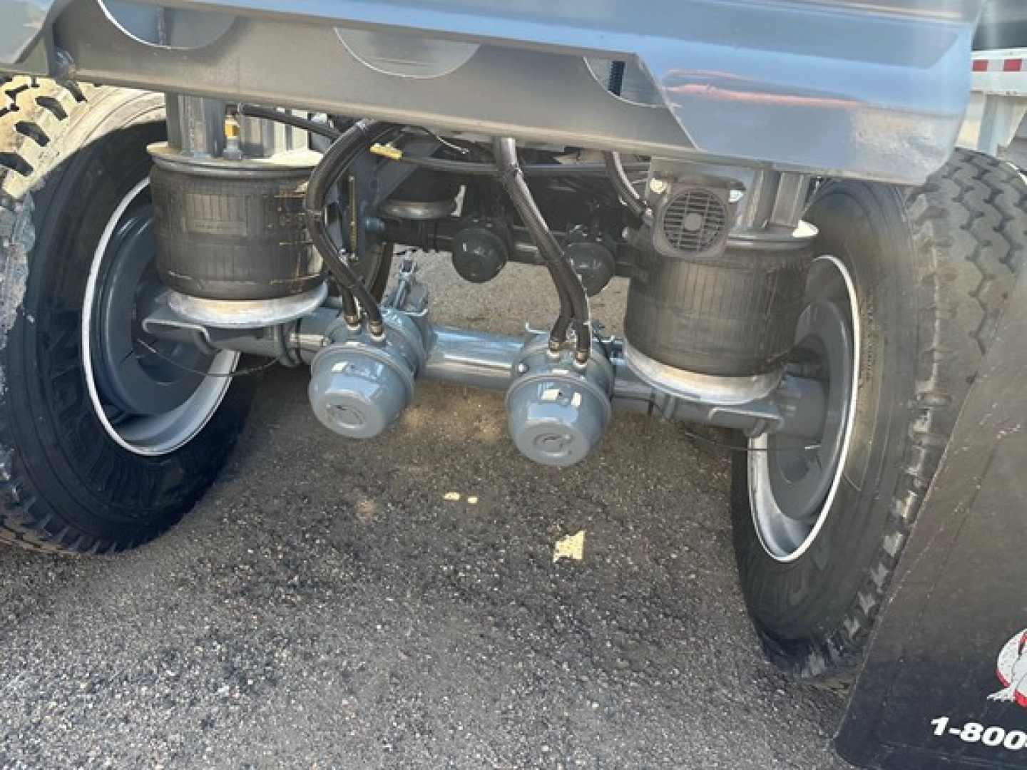 Trailer underside showing three gray hydraulic couplers and lines, two large wheels, and suspension components under a blue dump trailer.