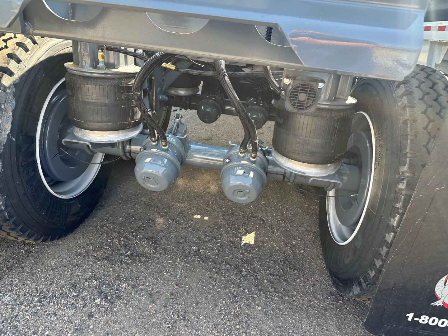 Underbody view of a trailer showing dual air or hydraulic lines connected to two round reservoirs and fittings between the wheels.