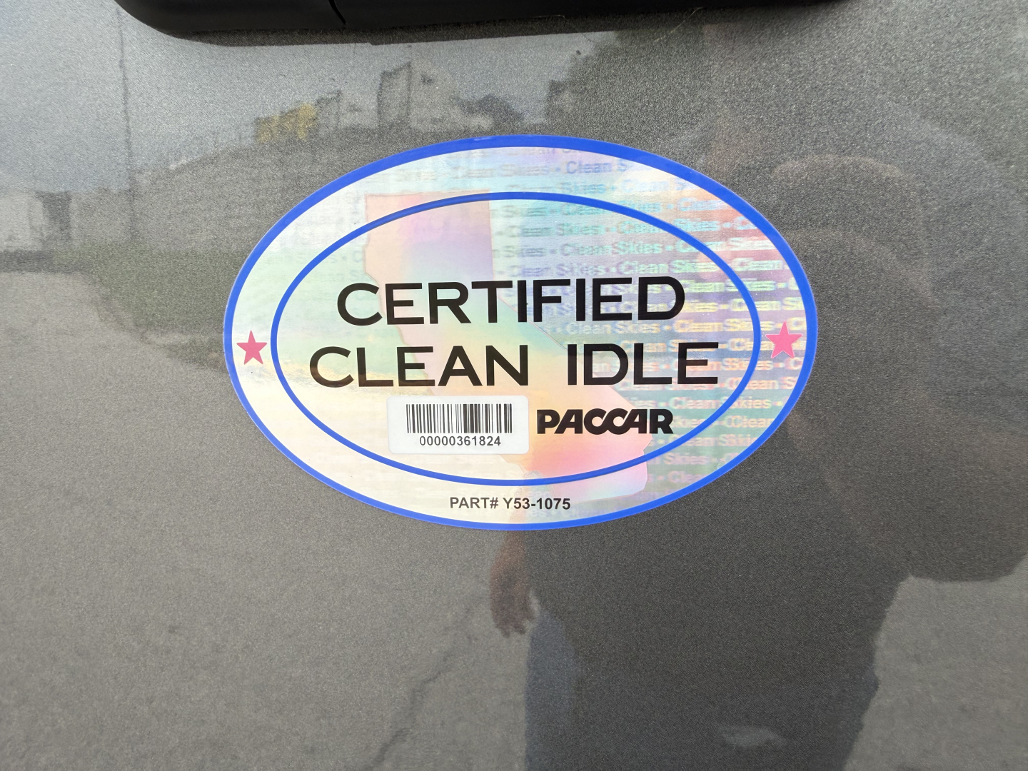 Sticker on vehicle showing CERTIFIED CLEAN IDLE with PacCAR logo and barcode, over reflection of person taking photo.