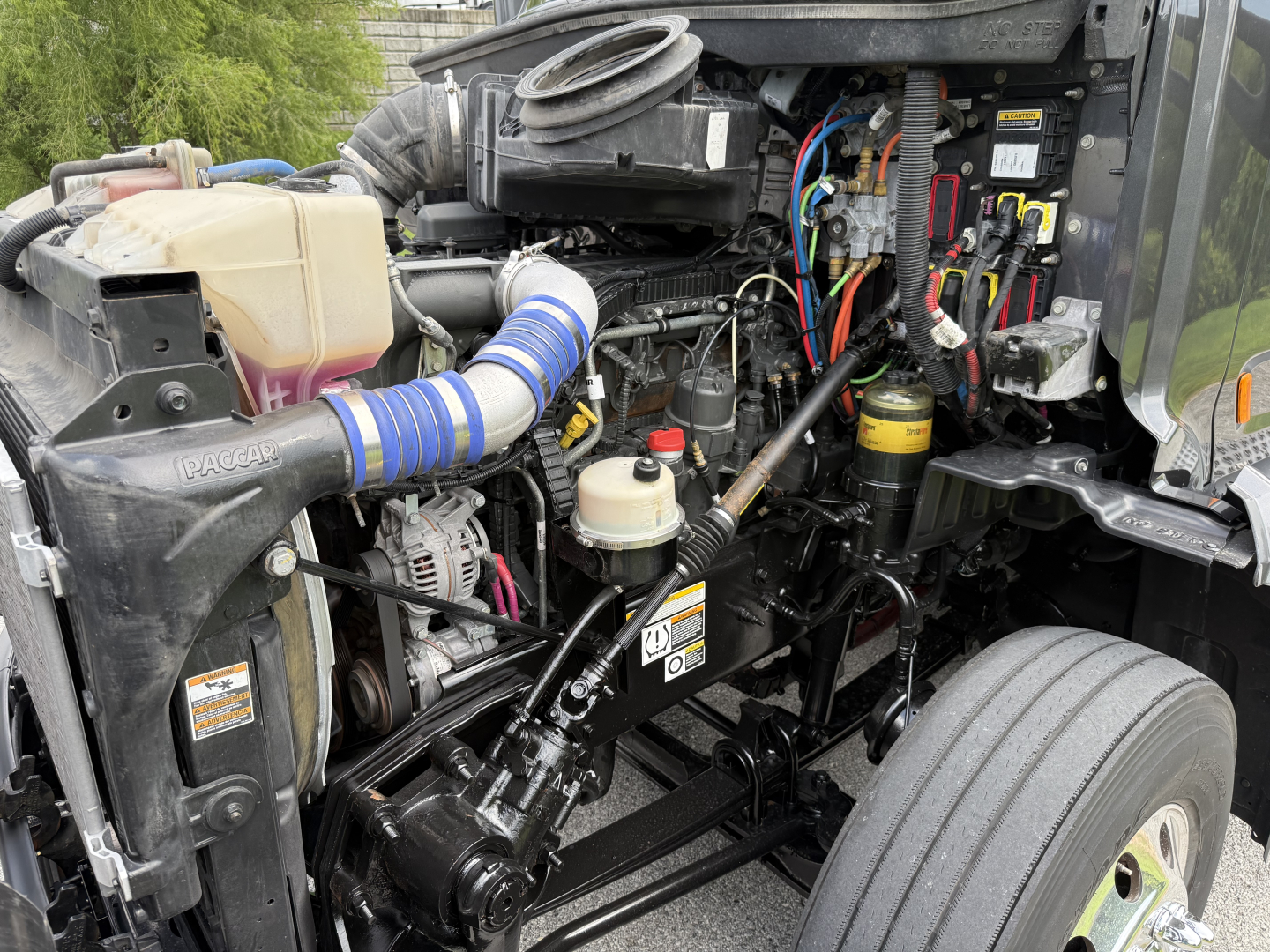 Peterbilt 579 engine bay showing front intake, coolant reservoir, hoses, wiring harnesses, and battery area.