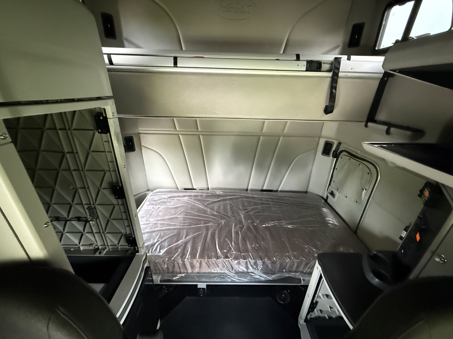 Interior of a 2022 Peterbilt 579 sleeper cab showing a diagonal storage shelf, side cabinets, and a plastic-covered bed in the sleeper area.