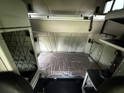 Interior of a 2022 Peterbilt 579 sleeper cab showing a diagonal storage shelf, side cabinets, and a plastic-covered bed in the sleeper area.