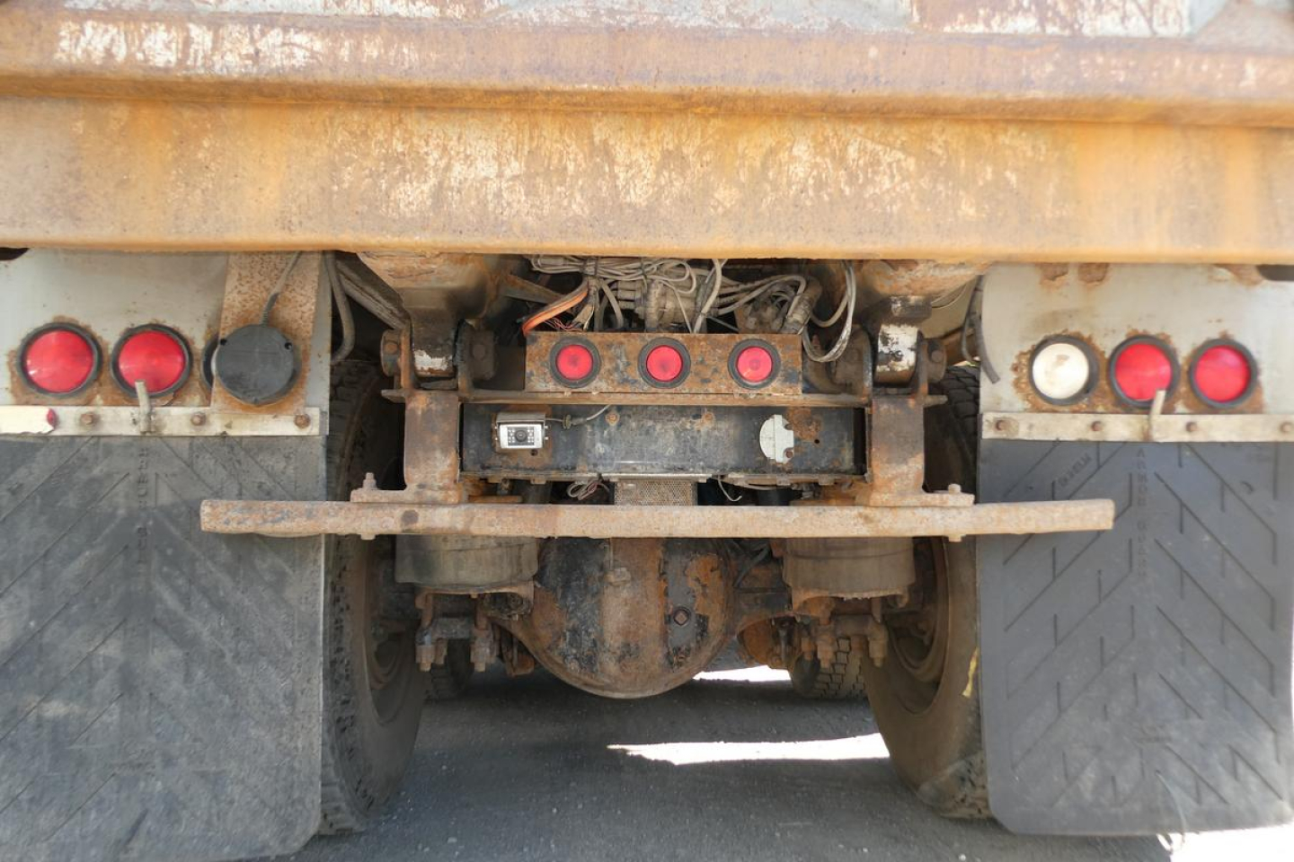 Rear view of a dump truck chassis showing bumper with taillights and hitch, rusted frame, and differential undercarriage.
