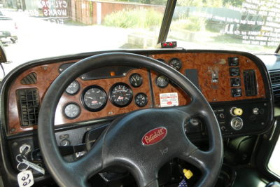 Peterbilt truck interior dashboard view with wood grain finish, steering wheel, and multiple gauges. Visible text includes 'Peterbilt' on the steering wheel center badge and various dash instruments.