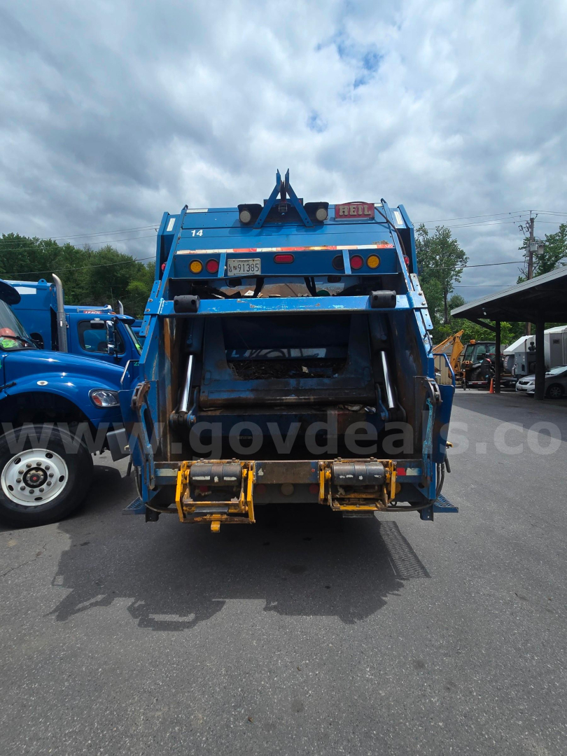 Back view of a blue Freightliner M2 106 garbage truck with a rear loader and hydraulic arms. Visible text: VIN plate area, stock number label, yellow hooks, and rear bumper.