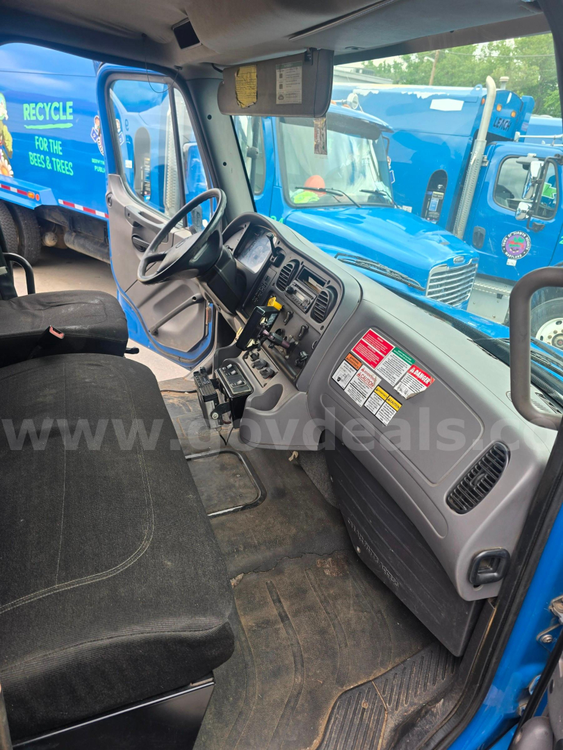 Cab interior of a 2012 Freightliner M2 106 truck showing steering wheel, dashboard with warning stickers, and front seats. Visible text includes safety/labels on the passenger side dash and instrument panel; background shows blue trucks out