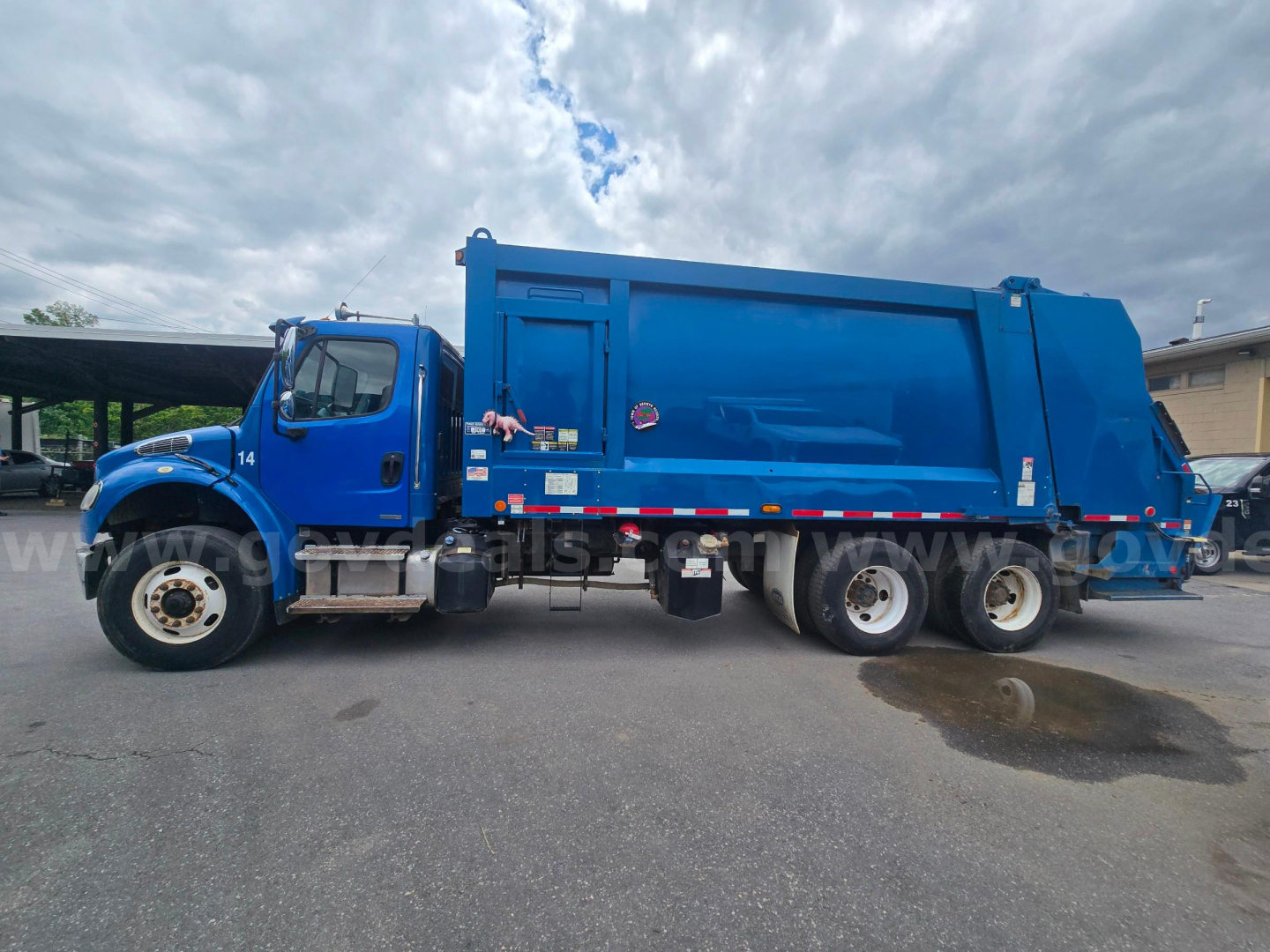 Garbage truck side view showing Freightliner M2 106 with blue body and rear refuse container. Visible cab, wheels, and decals along the side.