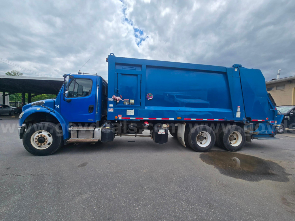 Garbage truck side view showing Freightliner M2 106 with blue body and rear refuse container. Visible cab, wheels, and decals along the side.