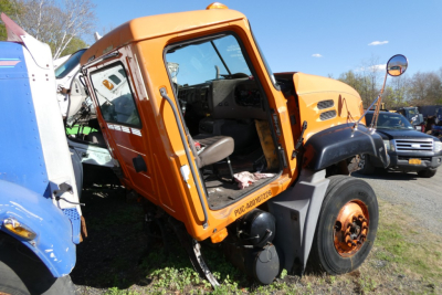 Orange Mack CX612 cab-end showing steering wheel and interior door; front wheel with hub; visible VIN plate area; outdoor truck lot.