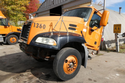Cab-over style Mack CX612 truck with orange cab and black fender, front left wheel raised, number 1360 on hood, side mirrors, and warning sign in background.