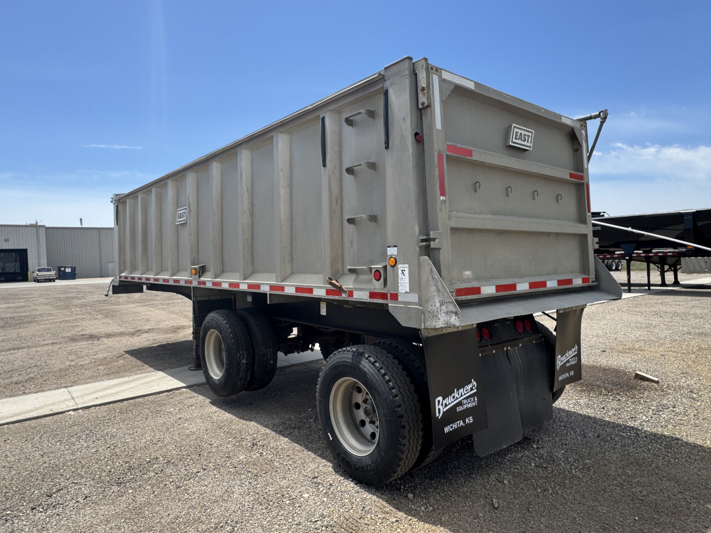 Dump trailer with dual rear axles and tall metal bed, gray in color, rear tailgate, reflective striping, and company mud flaps.