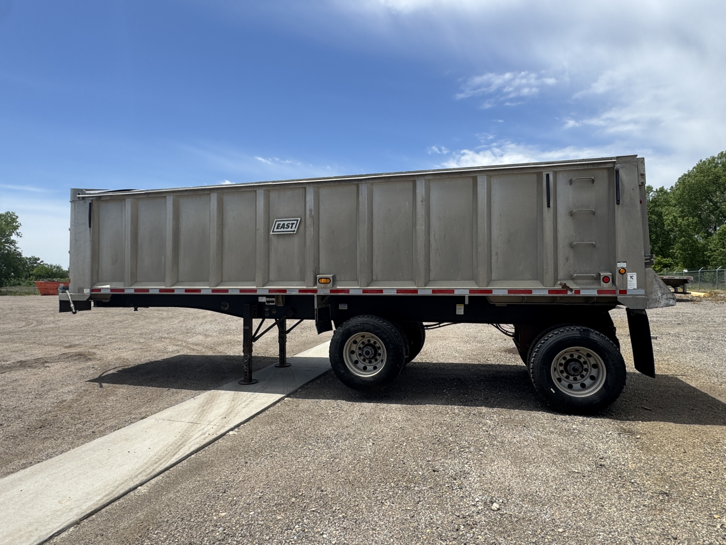 Dump trailer in side view showing dual tandem wheels and metal cargo bed with reflective tape and support legs.