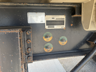 Diesel trailer hitch area with labels and stickers. Shows VIN 1E1D1L2876RC38818 and various circular safety decals; rectangular certification label and a smaller white label. Shows metal frame with bolts.
