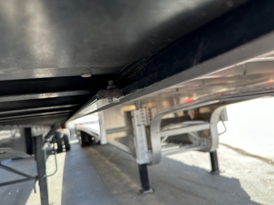 Trailer underside view showing frame, support legs, and hitch area with metal components and reflective surfaces, from a low angle under a flatbed trailer.