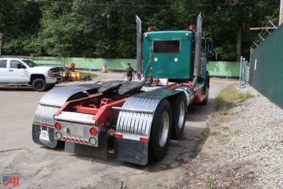 Kenworth W900 truck tractor rear view showing drivetrain and chrome fenders, green cab, fifth wheel, and dual rear axles.