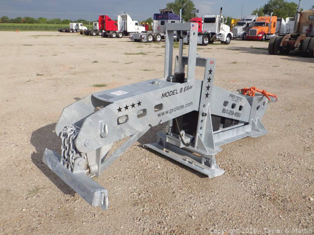 Pro-Tote 8 Elite hydraulic fifth wheel wrecker on a gravel lot, metal silver frame with hydraulic components, model 8 Elite text visible, chain and hitch hardware, several trucks in background.