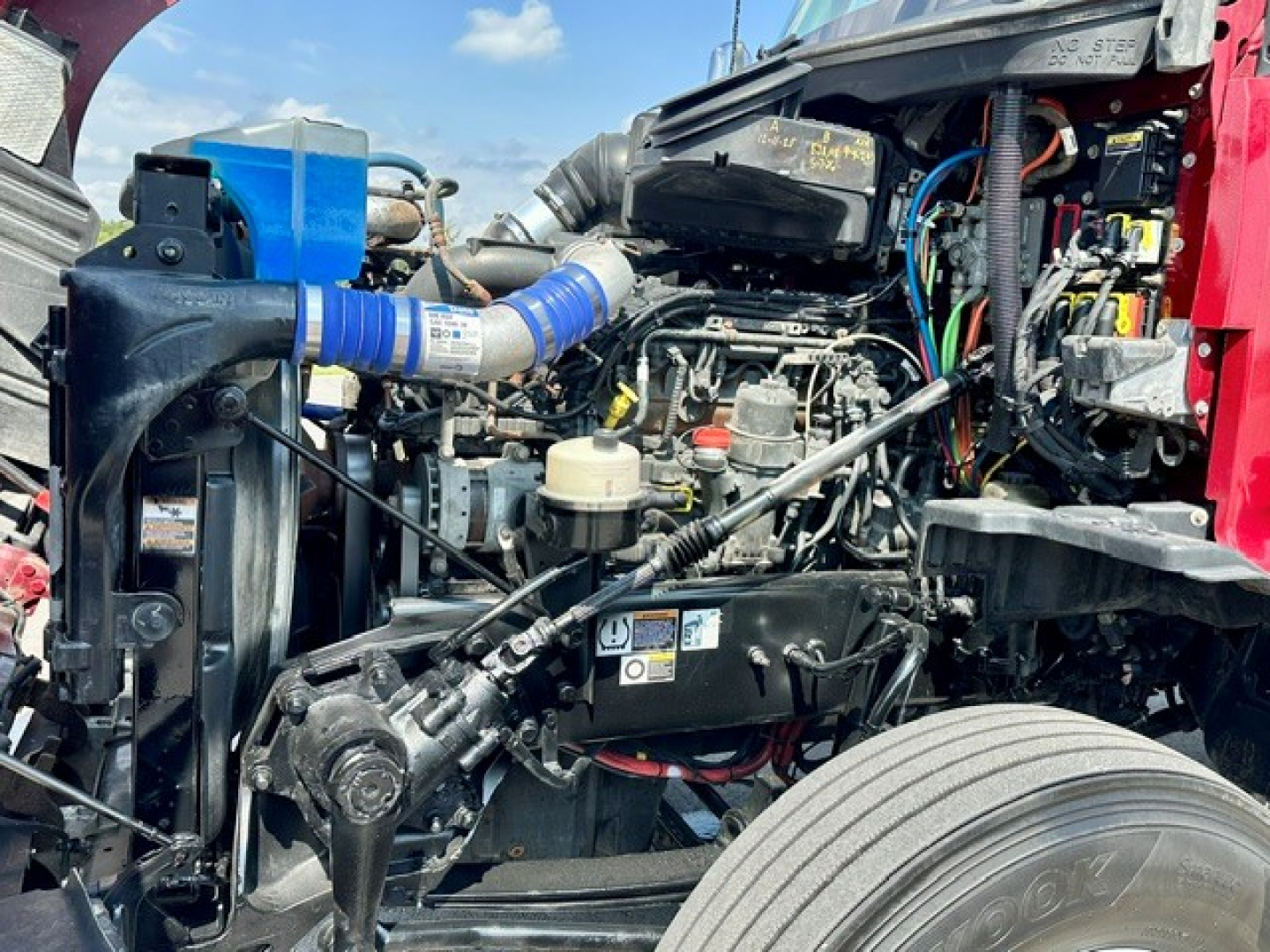 Kenworth T680 engine bay visible with interconnected hoses, wiring, and components including battery, coolant reservoir, and air intake piping.